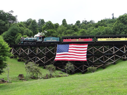 Locomotive No. 190 pulling passenger cars across the trestle at Tweetsie Railroad with a large American flag during the July 4 celebration